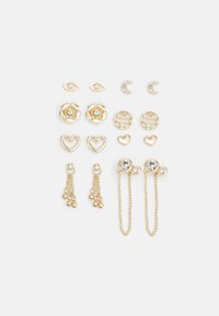 Conjunto de pendientes en tono dorado con diversas formas: flores, corazones, ojos, lunas y cadenas; algunos con cristales claros; diseños y texturas mezclados.