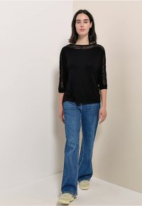 Top nero con maniche in pizzo a tre quarti, abbinato a jeans svasati di un blu chiaro. La modella si trova di fronte a uno sfondo neutro, indossando sneakers chiare.