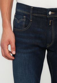 Tumedat sinised denim teksad, millel on kollane õmblus, viie-tasku disain ja peen tekstuur. Lähedalt vaadatuna on näha vöökohaga ja taskute detaile.