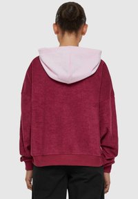 Bordeaux hoodie met een roze binnenkap, zachte textuur, relaxte pasvorm en geribbelde manchetten, van achteren gezien tegen een neutrale achtergrond.