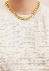 Złoty naszyjnik z ogniwami połączonymi, położony na teksturowanym, jasnym swetrze z dzianiny, który ma wzór w kratkę.