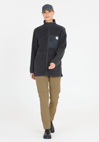 Veste en polaire gris foncé avec fermeture éclair à l'avant, deux grandes poches avant et une poche sur la poitrine. Associée à un pantalon beige et des bottes noires.