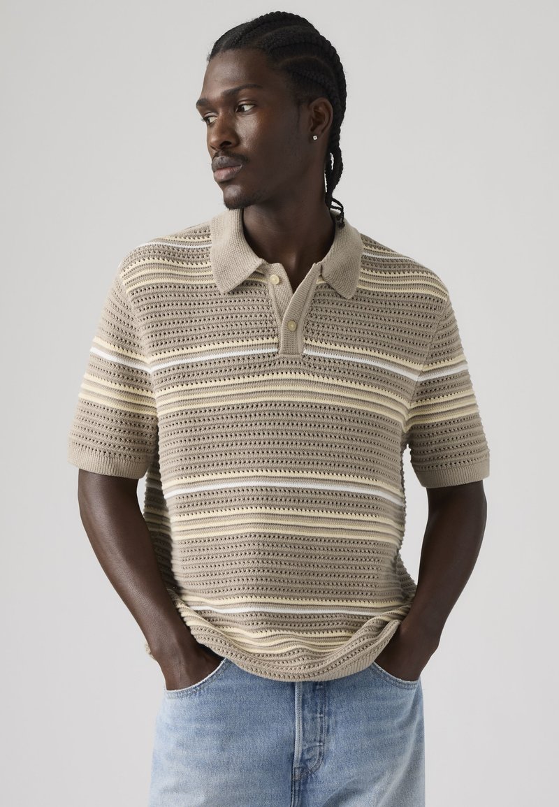 Homme avec des cheveux tressés portant un polo en tricot beige à rayures et un jean bleu clair, les mains dans les poches, regardant vers la gauche.