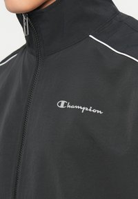 Črna jopica z zadrgo in visokim ovratnikom, z belimi detajli in srebrnim logotipom "Champion" na prsih. Gladka tekstura blaga.