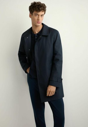 Młody mężczyzna w ciemnogranatowym płaszczu nałożonym na czarną koszulę, stojący z jedną ręką w kieszeni na jasnym, jednolitym tle.