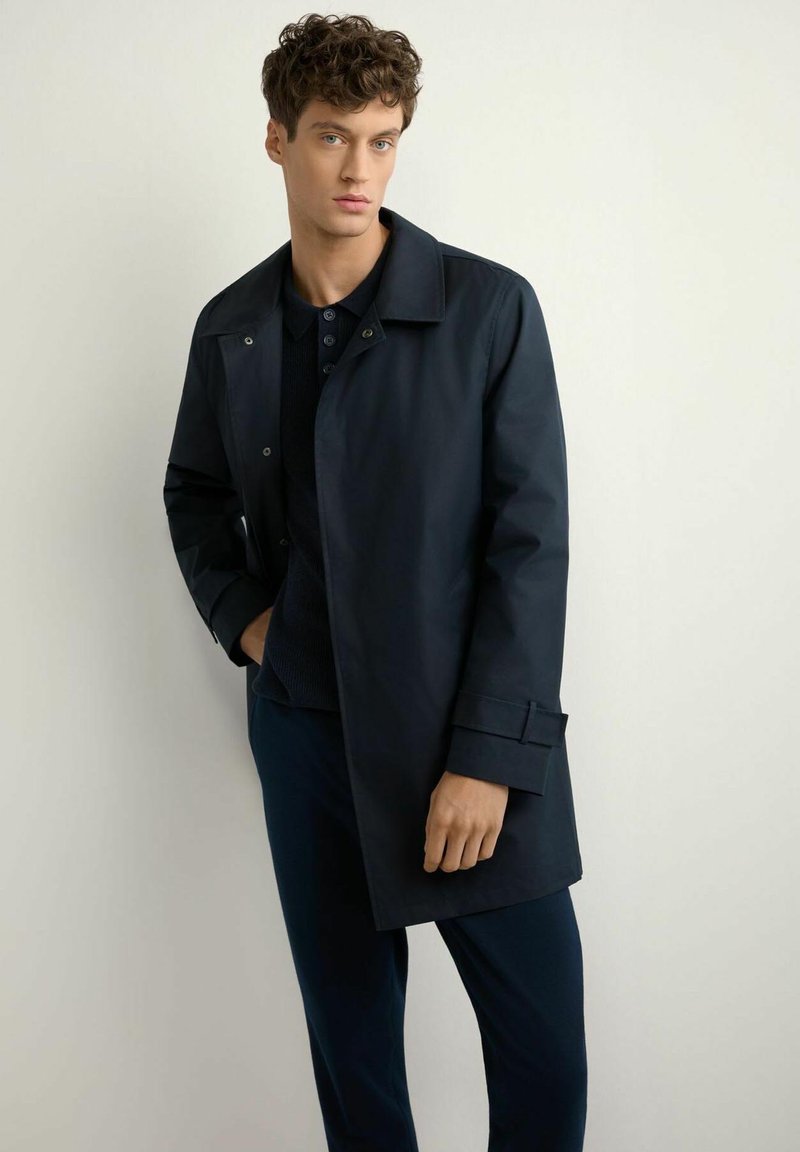 Młody mężczyzna w ciemnogranatowym płaszczu nałożonym na czarną koszulę, stojący z jedną ręką w kieszeni na jasnym, jednolitym tle.