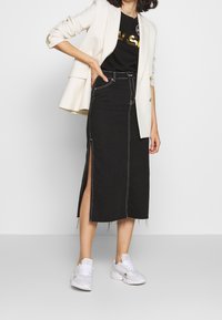 Falda midi de mezclilla negra con abertura lateral, diseño de cintura alta, costuras blancas, combinada con una camiseta gráfica negra y una blazer color crema.