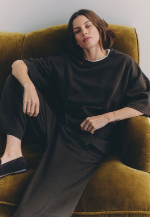 Femeie cu păr castaniu, îmbrăcată într-o ținută neagră supradimensionată, stând relaxată într-un fotoliu tapitat cu catifea muștar, uitându-se către cameră cu o expresie neutră.