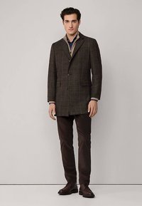Bruine ruitblazer met een geknotte kraag, twee knopen en een lange pasvorm. Gecombineerd met donkere broek en bruine schoenen. Gladde stoftextuur.