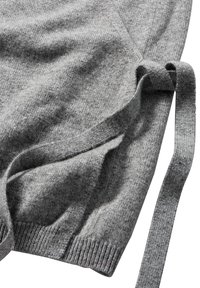 Hellgrauer Pullover mit weicher Textur, langen Bändern an der Seite und gerippten Bündchen. Der Stoff wirkt gemütlich und dehnbar.