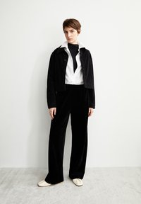Schwarze Cordjacke über einem weißen Hemd, kombiniert mit schwarzen Samt-Hosen mit weitem Bein und beigen Schuhen. Minimalistisches Design, weiche Texturen.