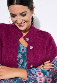 Fuchsia gestrickte Strickjacke mit geripptem Kragen und silbernen Knopfakzenten, kombiniert mit einem blumengemusterten Kleid in Petrol- und Rosatönen.