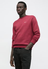 Felpa bordeaux con maniche lunghe e scollo a girocollo, caratterizzata da un piccolo logo bianco sul petto. Abbinata a jeans neri.