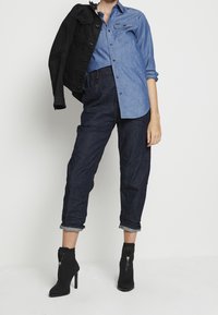 Person med uppvikta mörka jeans, blå skjorta med knappar, svart jacka hängande över en axel och svarta hälsäkta ankelstövlar.