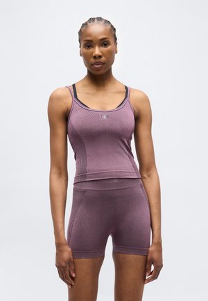 Donna con capelli intrecciati che indossa un top senza cuciture e pantaloncini abbinati color malva, in piedi davanti a uno sfondo chiaro e uniforme.