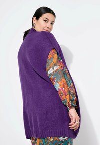 Lila gestrickte Weste mit strukturiertem Finish, mit offenem Vorderteil und Seitenschlitzen, über einem bunten, floral gemusterten Langarmhemd.