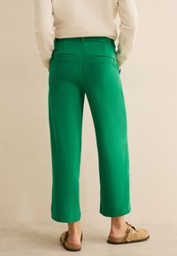 Pantalon vert à jambes droites avec une texture lisse, taille élastique et deux poches arrière, associé à des mocassins marron clair.