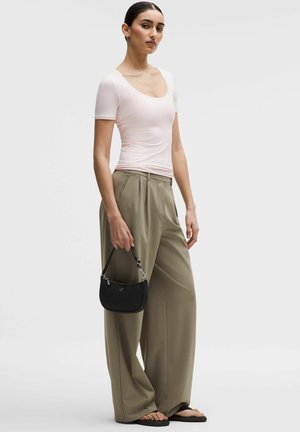 Frau trägt ein hellrosa eng anliegendes Kurzarmoberteil, weit geschnittene khakifarbene Hose, schwarze Sandalen und hält eine kleine schwarze Handtasche seitlich.