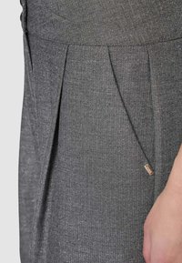 Primo piano di pantaloni in tessuto grigio spina di pesce con pieghe, tasche e una piccola targhetta metallica visibile vicino alla mano di una persona.