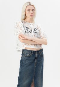 Blouse courte en dentelle blanche avec des manches bouffantes, ornée de motifs floraux, associée à une jupe en denim taille haute avec une fente à l'avant.