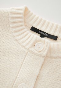 Cremefarbener, gestrickter Pullover mit einem gerippten Kragen, zwei weißen Knöpfen und einem schwarzen Etikett mit dem Markennamen "someday."
