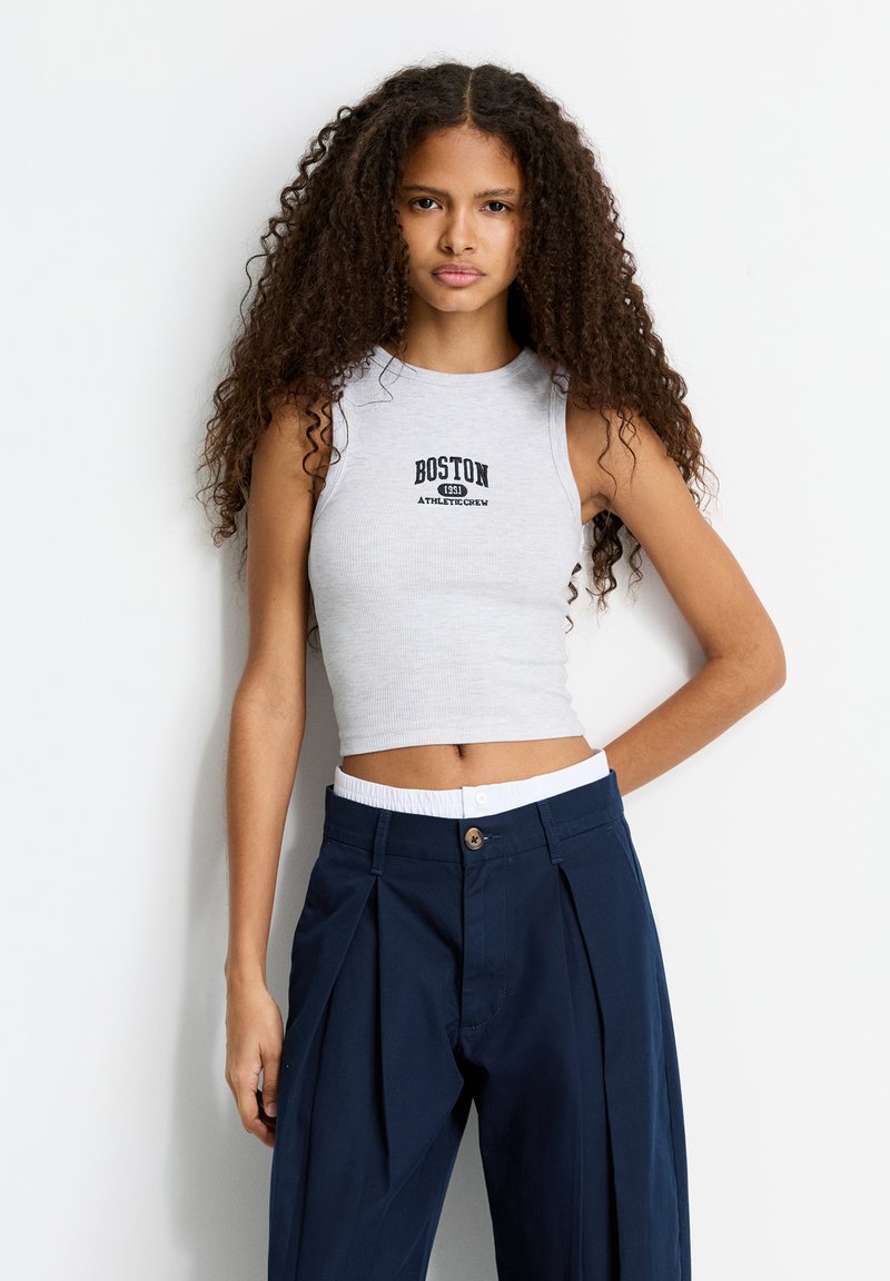 Šedý rebrovaný crop top so znakmi "BOSTON 1953 ATHLETIC CREW", kombinovaný s námorníckymi plisovanými nohavicami s bielym elastickým pásom.