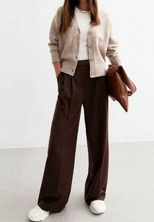 Žena v béžovém kardiganu, bílém topu, širokých tmavě hnědých kalhotách, bílých teniskách, držící velkou hnědou kabelku s texturou.