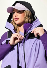 Donna che indossa una giacca con cappuccio lavanda e nera e un berretto coordinato lavanda, mentre chiude il collo della giacca.