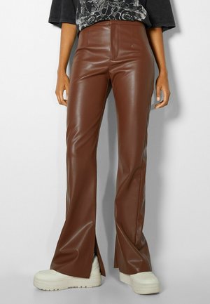 Persoon draagt hooggetailleerde bruine nepleren broek met vooropeningen, gecombineerd met witte chunky sneakers en een zwart grafisch t-shirt.