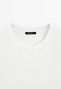 Bílé žebrované tričko s kulatým výstřihem a hladkým povrchem. Štítek uvádí "Massimo Dutti, vyrobeno v Maroku" připevněný na krku.