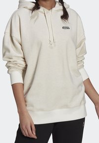 Osoba v krémové mikině Adidas s kapucí a malým logem na hrudi, v kombinaci s černými kalhotami.