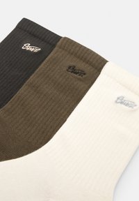 Tre par af ribbede sokker i mørkegrå, olivengrøn og creme. Hver har et lille mærkelogo broderet nær kanten.