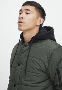 Grønn isolert jakke med quiltet design, med svarte detaljer, hette, knapper og en glidelåslomme på ermet.