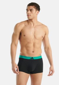 Boxer-briefs noirs pour hommes avec une ceinture élastique verte arborant un logo. Le tissu semble lisse et extensible, conçu pour le confort.