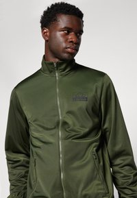 Groene sportjack met een hoge kraag, ritssluiting aan de voorkant en zijzakken. Bevat een logo op de borst en heeft een gladde, glanzende textuur.