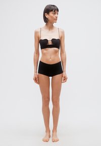 Γυναίκα όρθια ξυπόλητη με μαύρα εσώρουχα δύο τεμαχίων, κοιτάζοντας προς τα πλάγια σε απλό ανοιχτό φόντο.