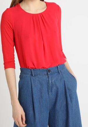 Femme portant un chemisier rouge vif à manches longues, rentré dans un pantalon en jean bleu taille haute, ample, avec des poches.