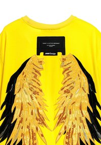 Camiseta amarilla con intrincados diseños de alas bordados en negro y dorado. Incluye una etiqueta rectangular con información de la marca en el cuello.