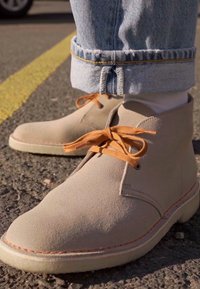 Botas de tobillo de gamuza beige con cordones naranjas, puntera redonda y costuras en contraste. Llevadas con jeans de denim arremangados, de pie sobre una superficie de asfalto.