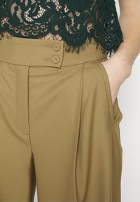 Olijfgroene broek met een gladde textuur, voorzien van een hoge taille, sluiting met twee knopen en zijzakken. Ook een kanten top is zichtbaar.