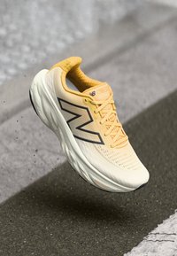 Běžecká obuv New Balance v světlé žluté barvě s texturovaným svrškem, černým logem a polstrovanou podrážkou, levitující nad betonovým povrchem.