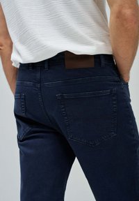Jeans de denim azul oscuro con una textura suave, que presentan una etiqueta de cuero marrón en la cintura y dos bolsillos posteriores con un sutil detalle de costura.