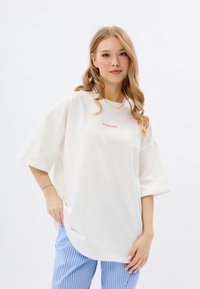 Witte oversized t-shirt met korte mouwen, met roze tekst "MIRONS" op de voorkant. Gecombineerd met lichtblauwe gestreepte broek.