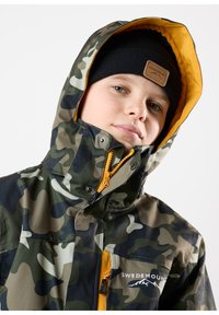 Kamouflagemönstrad jacka med dragkedja framtill och orange detaljer, med huva och en logotyp. Svart stickad mössa bärs under.