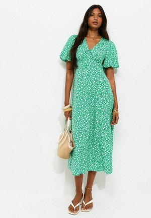 Femme portant une robe midi verte à fleurs avec manches bouffantes, tenant un sac à main beige tressé, et portant des sandales blanches et des bijoux dorés.
