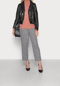 Giacca da biker in pelle nera su un dolcevita rosa, abbinata a pantaloni cropped grigi a scacchi con texture e tacchi a punta nera.