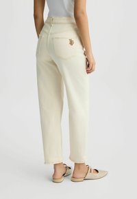 Jeans beige clair taille haute avec une coupe décontractée, arborant des détails en métal doré, des poches arrière et un ourlet effiloché. Associés à des sandales ornées.