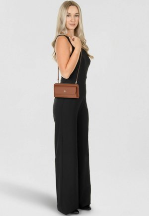 Frau mit langen blonden Haaren, die einen schwarzen ärmellosen Overall trägt und eine kleine braune Leder-Umhängetasche bei sich hat.