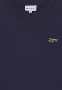 Felpa blu navy con collo a girocollo a costine, caratterizzata da un piccolo logo di coccodrillo verde e bianco sul lato sinistro del petto. Texture morbida e design semplice.