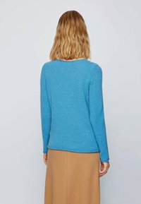 Femme aux cheveux blonds mi-longs portant un pull bleu à manches longues et une jupe couleur camel, de dos par rapport à la caméra.